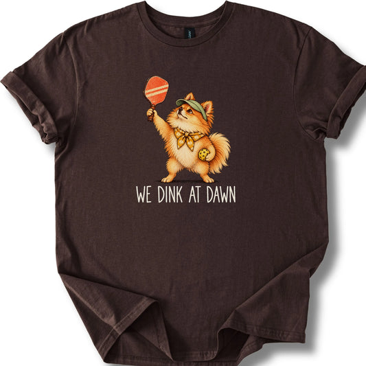 Pomeranian with a pickleball paddle and ball on a Dark Chocolate Gildan® T-shirt with We Dink at Dawn text
