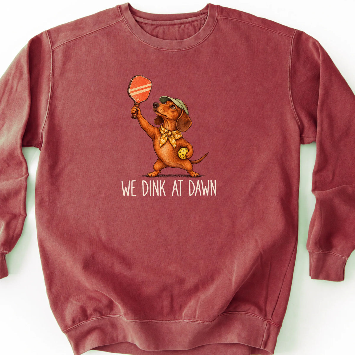 Dachshund with a pickleball paddle and ball on a Crimson Comfort Colors® sweatshirt with We Dink at Dawn text