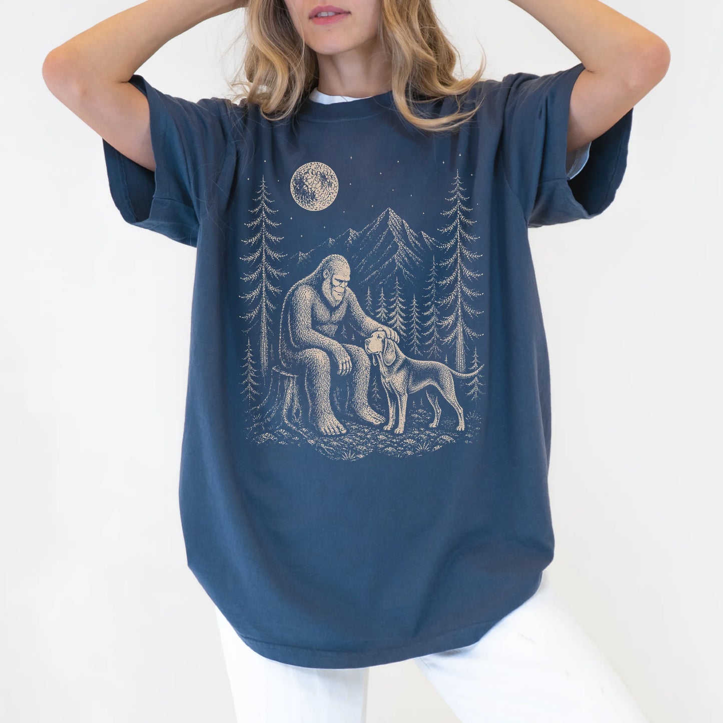 Model wearing a Denim Comfort Colors® T-shirt with bigfoot petting a bloodhound beneath the moon in a pine forest