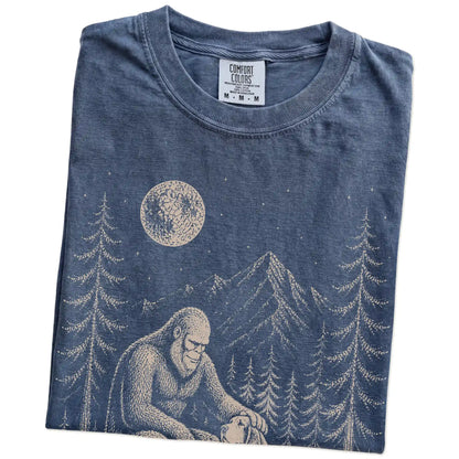 Folded Denim Comfort Colors® T-shirt showing bigfoot petting a bloodhound beneath the moon in a pine forest