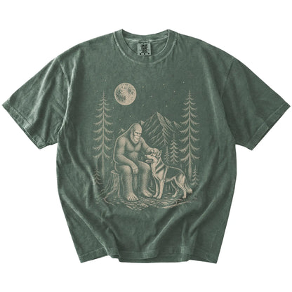 Bigfoot petting a German Shepherd beneath the moon in a pine forest on a Moss Comfort Colors® T-shirt