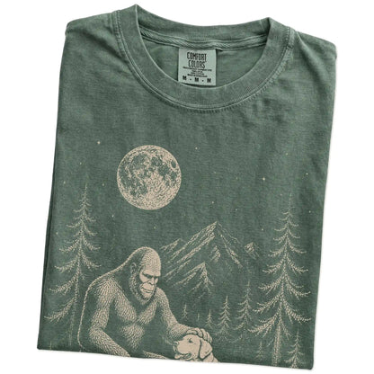 Folded Moss Comfort Colors® T-shirt showing bigfoot petting a labrador retriever beneath the moon in a pine forest