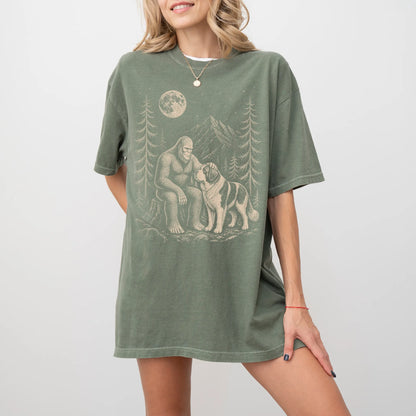 Model wearing a Moss Comfort Colors® T-shirt with bigfoot petting a saint bernard beneath the moon in a pine forest