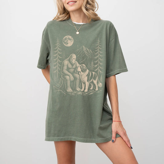 Model wearing a Moss Comfort Colors® T-shirt with bigfoot petting a saint bernard beneath the moon in a pine forest