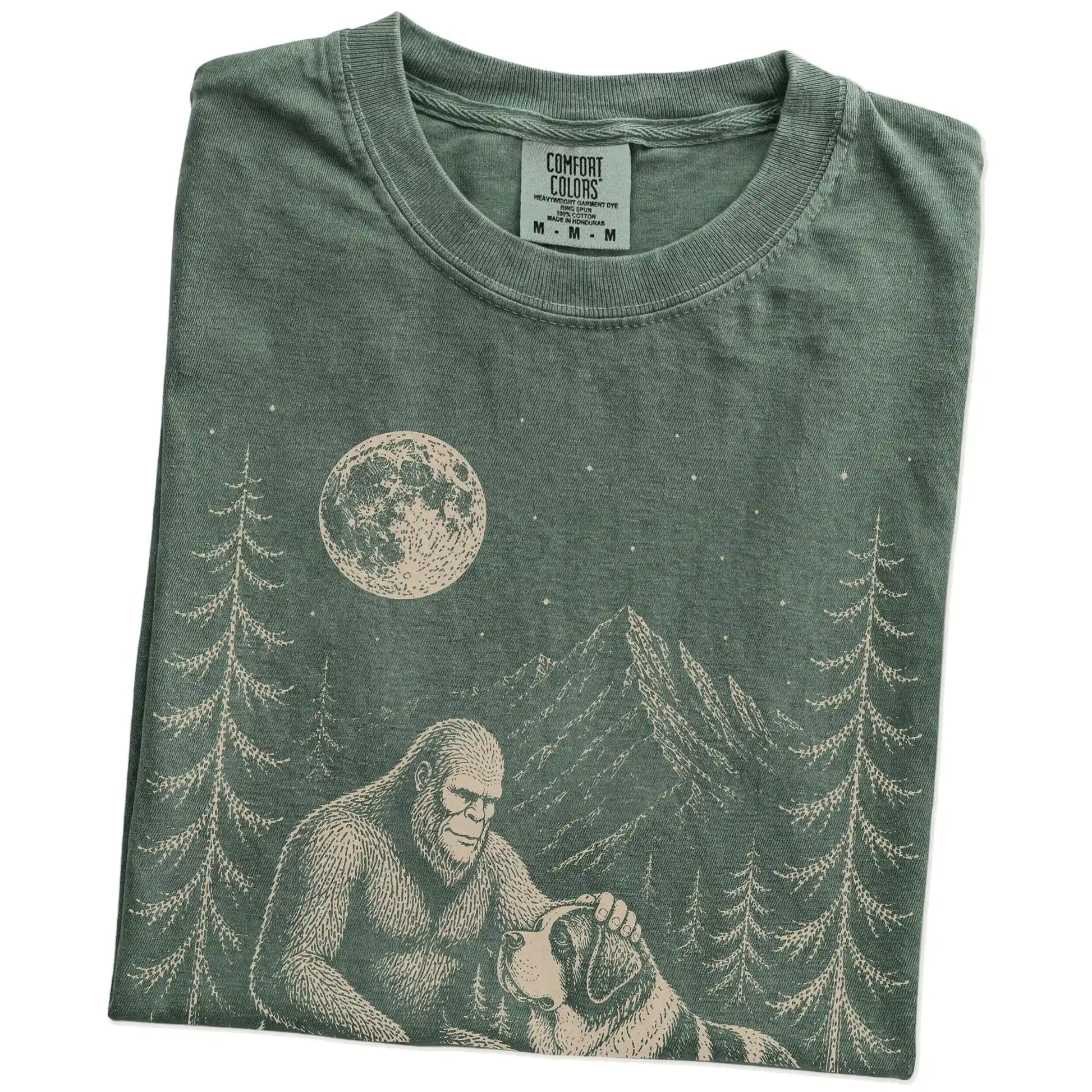 Folded Moss Comfort Colors® T-shirt showing bigfoot petting a saint bernard beneath the moon in a pine forest