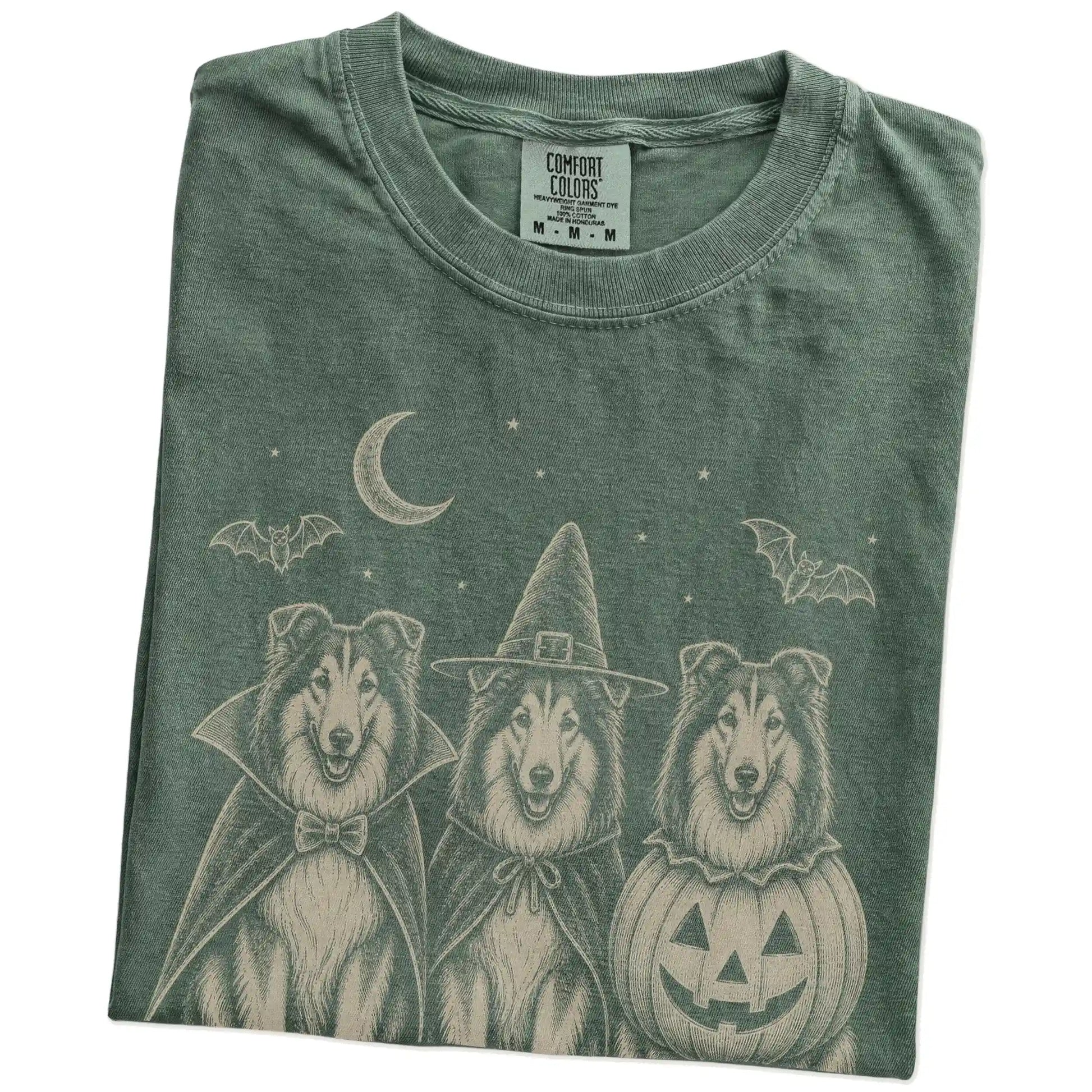 Folded Moss Comfort Colors® T-shirt showing three Collie dogs dressed for Halloween beneath bats and a crescent moon