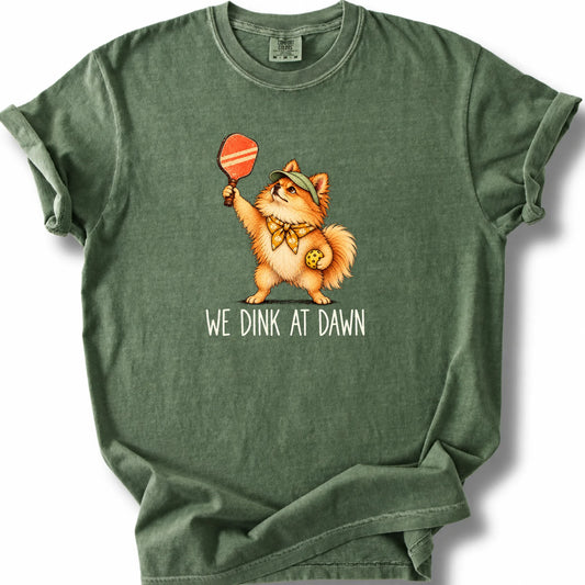 Pomeranian with a pickleball paddle and ball on a Moss Comfort Colors® T-shirt with We Dink at Dawn text