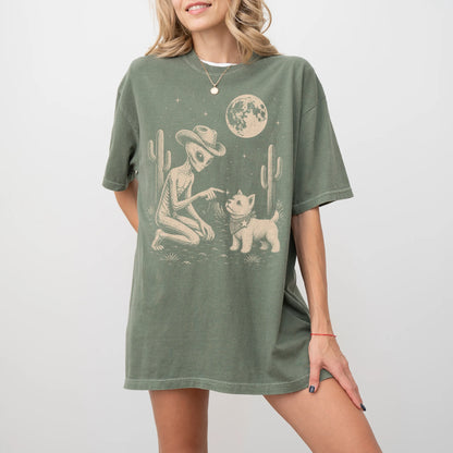 Model wearing a Moss Comfort Colors® T-shirt with cowboy alien fist-bumping a westie beneath the moon