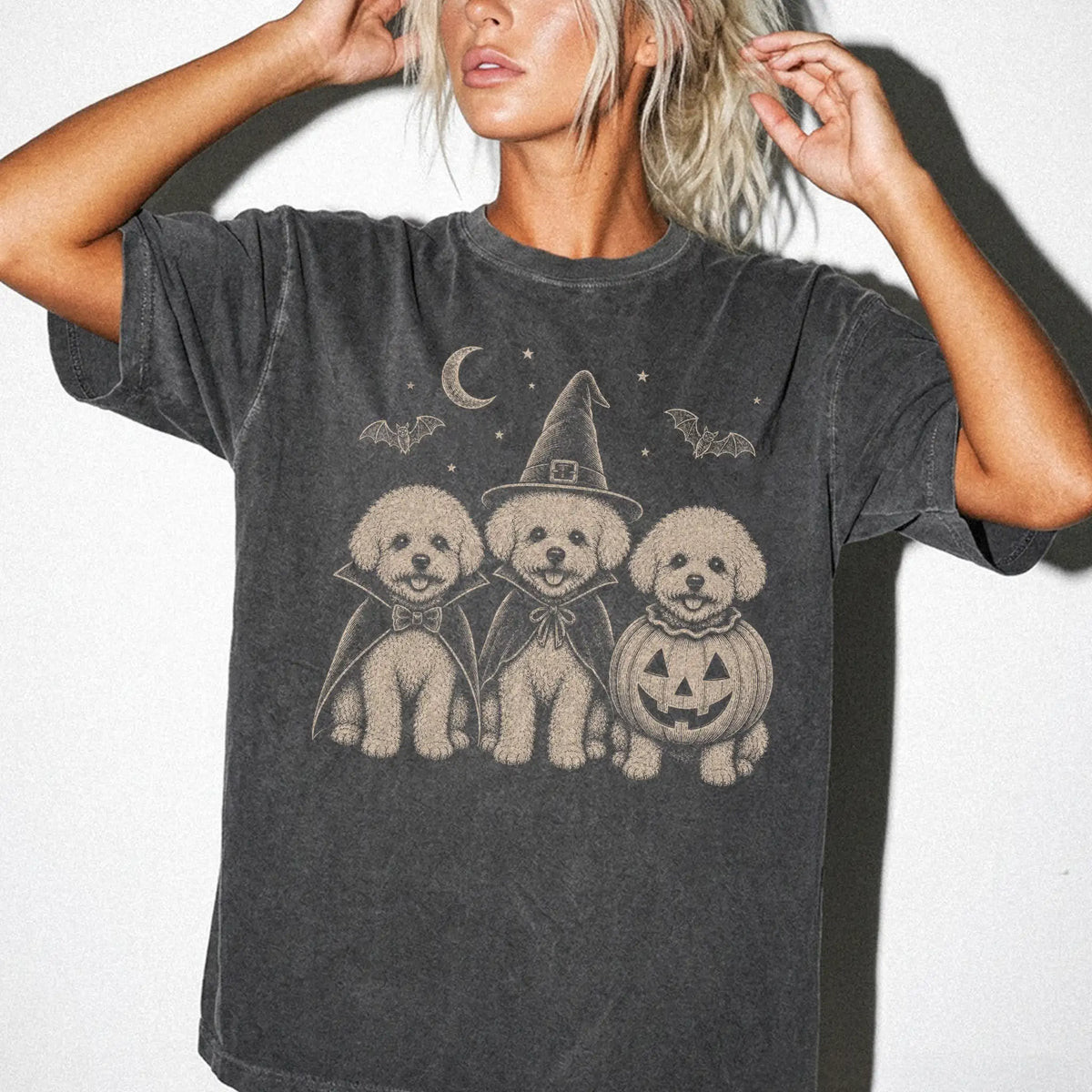 Model wearing a Pepper Comfort Colors® T-shirt showing three Bichon Frise dogs dressed for Halloween beneath bats and a cresc