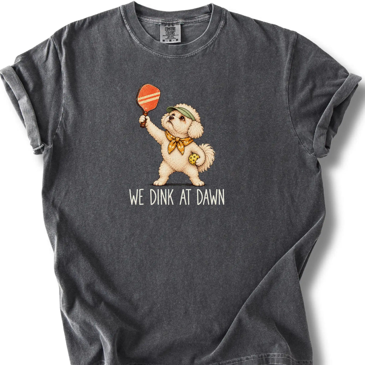 Bichon Frise with a pickleball paddle and ball on a Pepper Comfort Colors® T-shirt with We Dink at Dawn text
