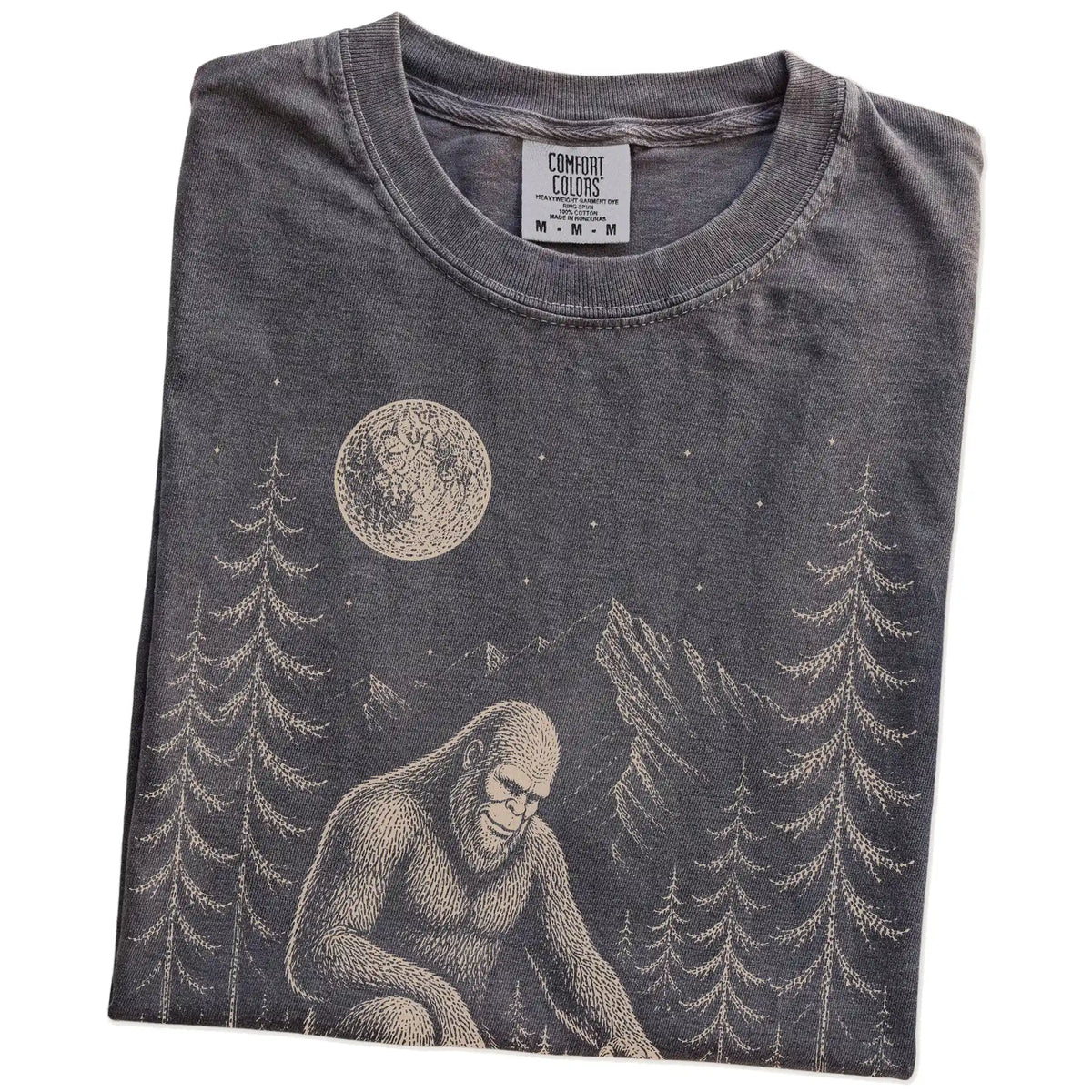 Folded Pepper Comfort Colors® T-shirt showing bigfoot petting a basset hound beneath the moon in a pine forest