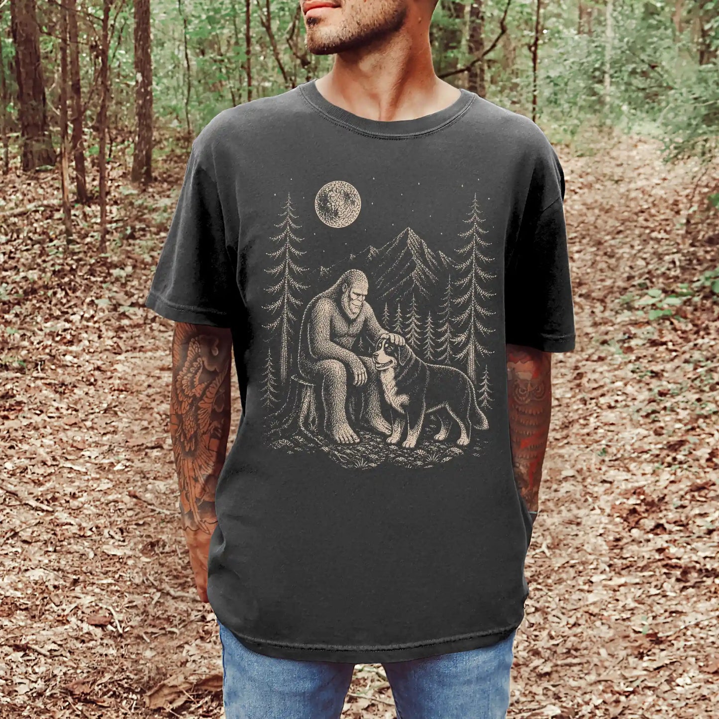 Model wearing a Pepper Comfort Colors® T-shirt with bigfoot petting a bernese mountain dog beneath the moon in a pine forest