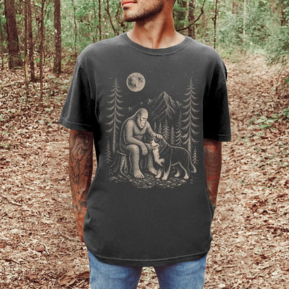 Model wearing a Pepper Comfort Colors® T-shirt with bigfoot petting a bernese mountain dog beneath the moon in a pine forest