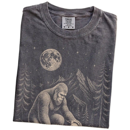 Folded Pepper Comfort Colors® T-shirt showing bigfoot petting a bichon frise beneath the moon in a pine forest