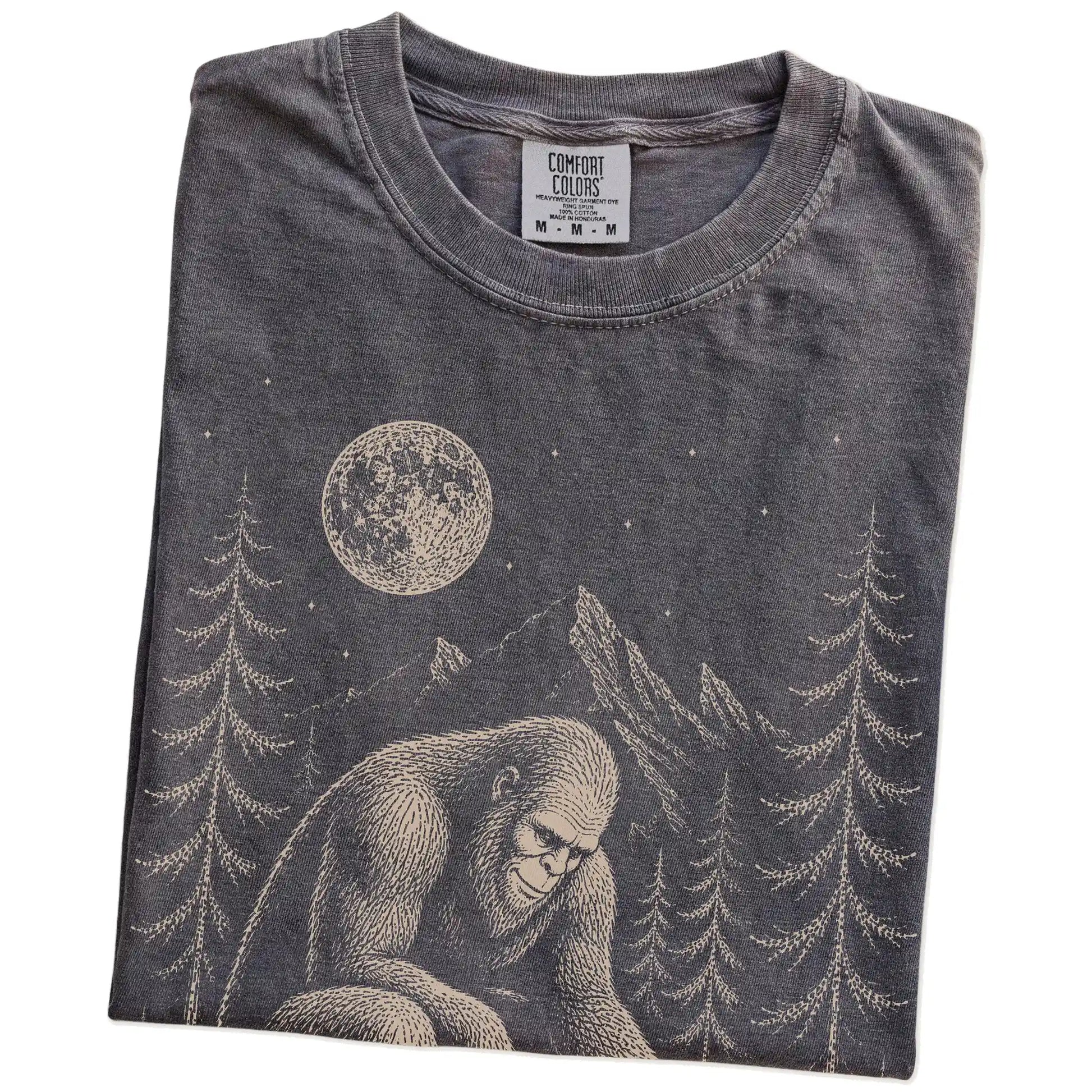 Folded Pepper Comfort Colors® T-shirt showing bigfoot petting a chihuahua beneath the moon in a pine forest