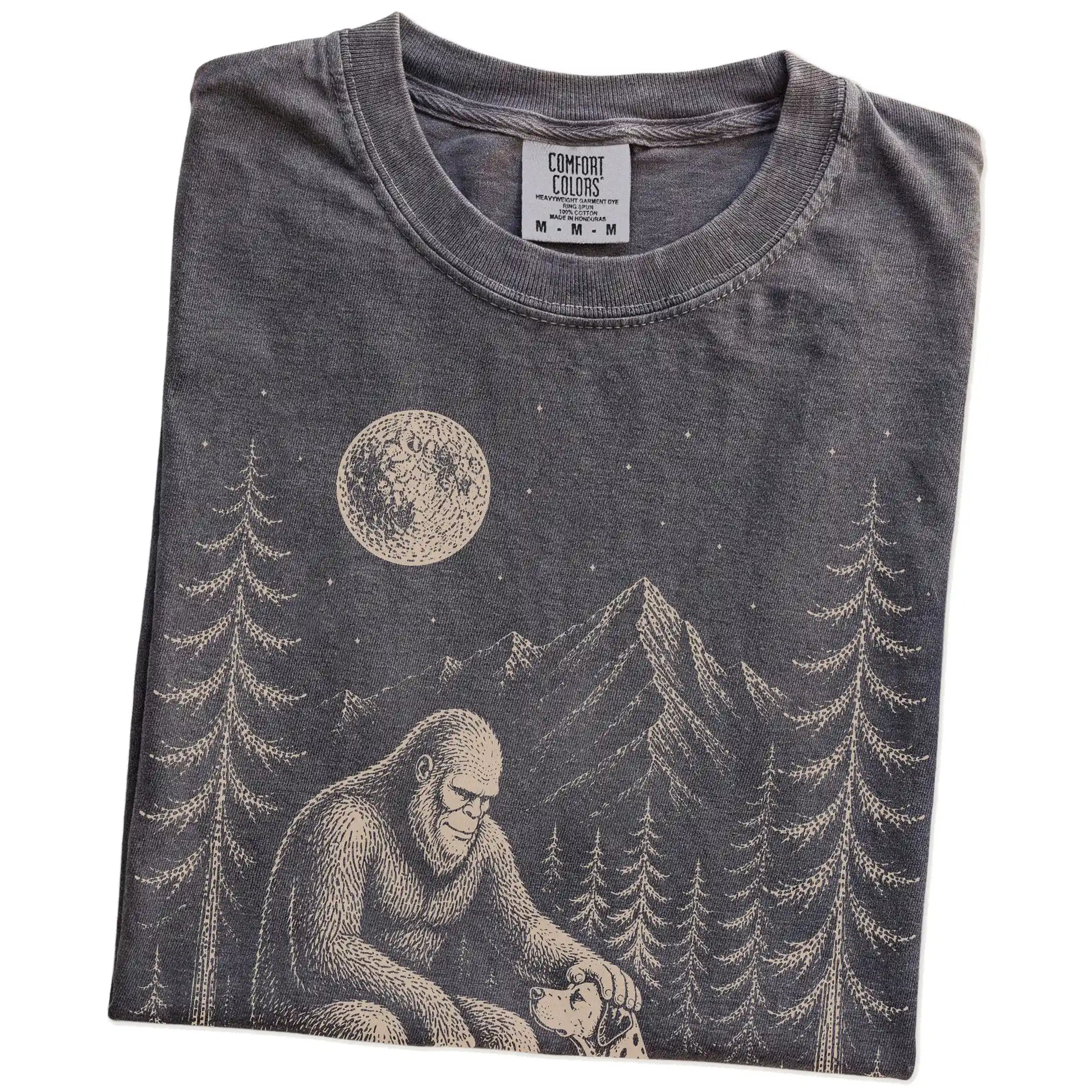 Folded Pepper Comfort Colors® T-shirt showing bigfoot petting a dalmatian beneath the moon in a pine forest
