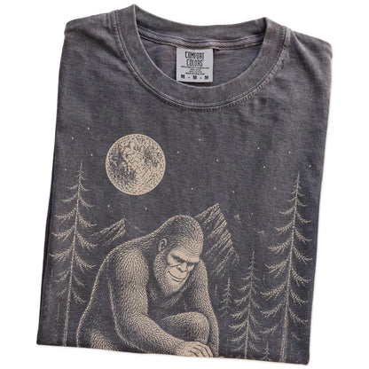 Folded Pepper Comfort Colors® T-shirt showing bigfoot petting a french bulldog beneath the moon in a pine forest