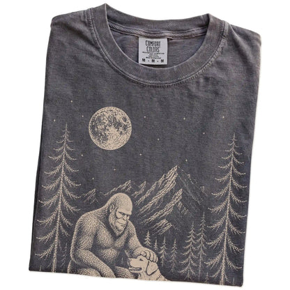 Folded Pepper Comfort Colors® T-shirt showing bigfoot petting a golden retriever beneath the moon in a pine forest