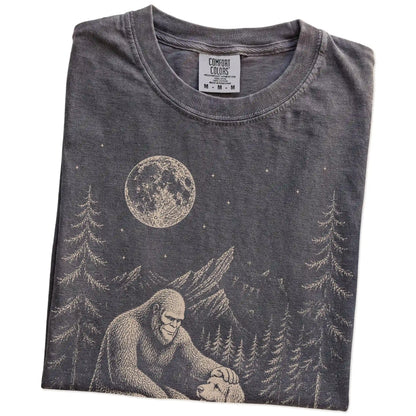 Folded Pepper Comfort Colors® T-shirt showing bigfoot petting a labradoodle beneath the moon in a pine forest