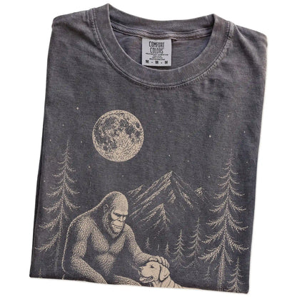 Folded Pepper Comfort Colors® T-shirt showing bigfoot petting a labrador retriever beneath the moon in a pine forest
