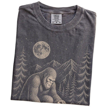 Folded Pepper Comfort Colors® T-shirt showing bigfoot petting a maltese beneath the moon in a pine forest