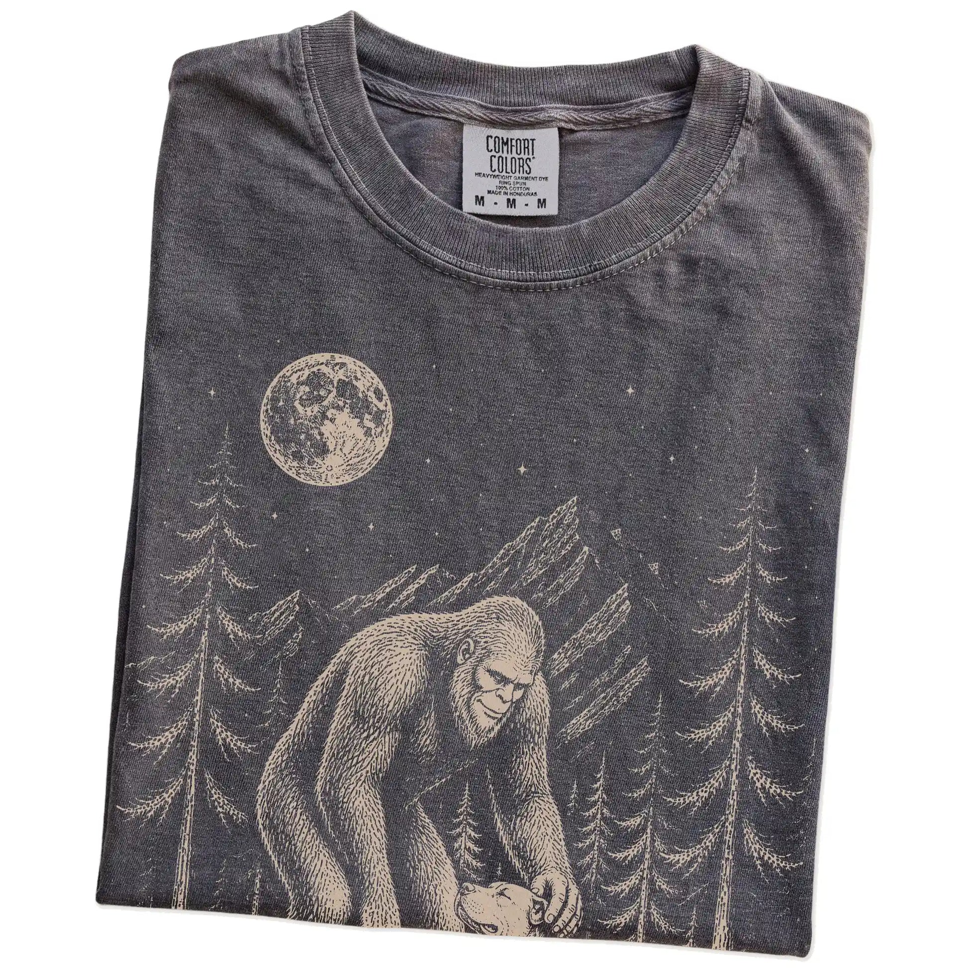 Folded Pepper Comfort Colors® T-shirt showing bigfoot petting a pitbull beneath the moon in a pine forest