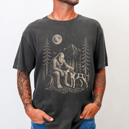 Model wearing a Pepper Comfort Colors® T-shirt with bigfoot petting a pointer beneath the moon in a pine forest