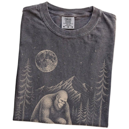 Folded Pepper Comfort Colors® T-shirt showing bigfoot petting a pomeranian beneath the moon in a pine forest