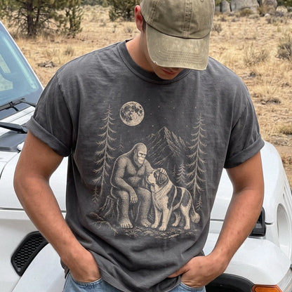 Model wearing a Pepper Comfort Colors® T-shirt with bigfoot petting a saint bernard beneath the moon in a pine forest