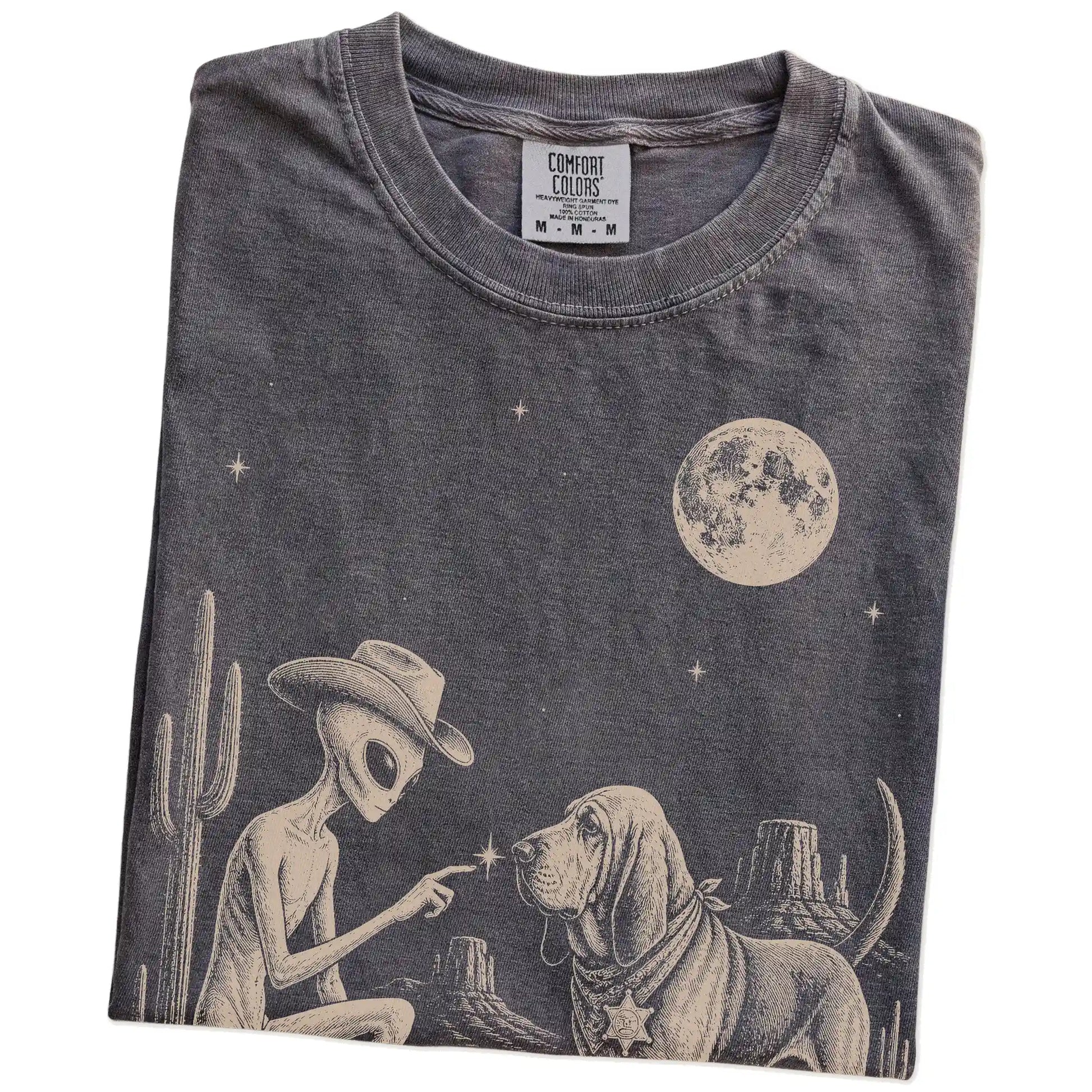 Folded Pepper Comfort Colors® T-shirt showing cowboy alien fist-bumping a bloodhound beneath the moon