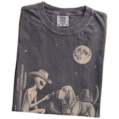 Folded Pepper Comfort Colors® T-shirt showing cowboy alien fist-bumping a bloodhound beneath the moon