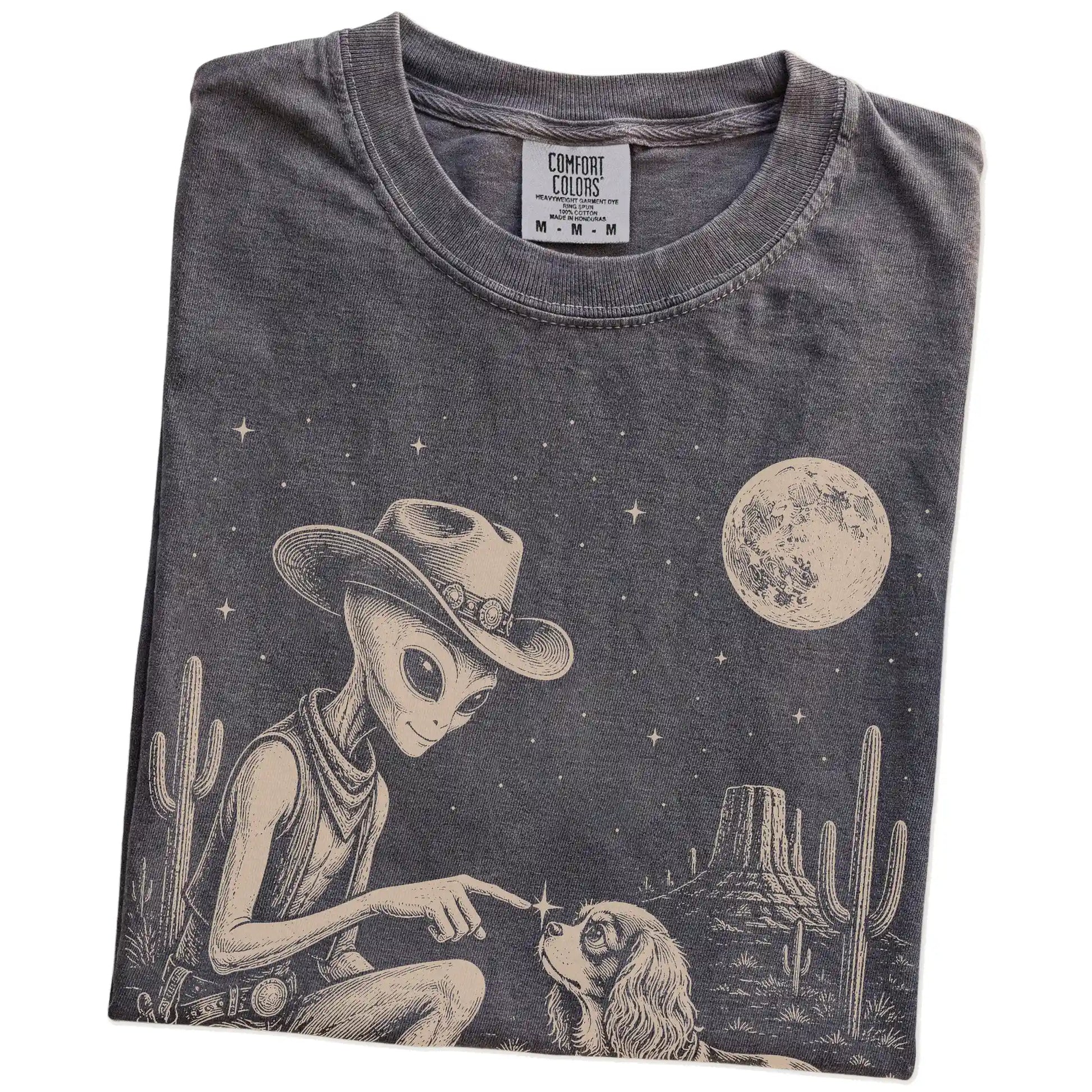 Folded Pepper Comfort Colors® T-shirt showing cowboy alien fist-bumping a cavalier king charles spaniel beneath the moon