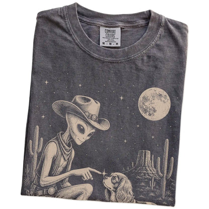 Folded Pepper Comfort Colors® T-shirt showing cowboy alien fist-bumping a cavalier king charles spaniel beneath the moon