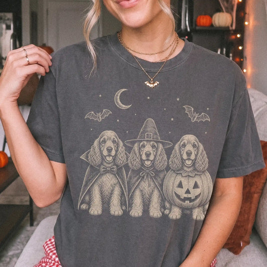 Model wearing a Pepper Comfort Colors® T-shirt showing three Cocker Spaniel dogs dressed for Halloween beneath bats and a cre
