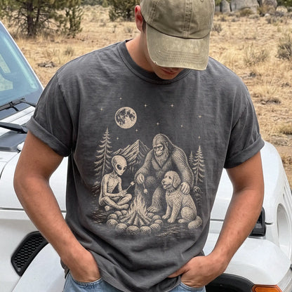Model wearing a Pepper Comfort Colors® T-shirt showing Goldendoodle camping with Bigfoot and an alien