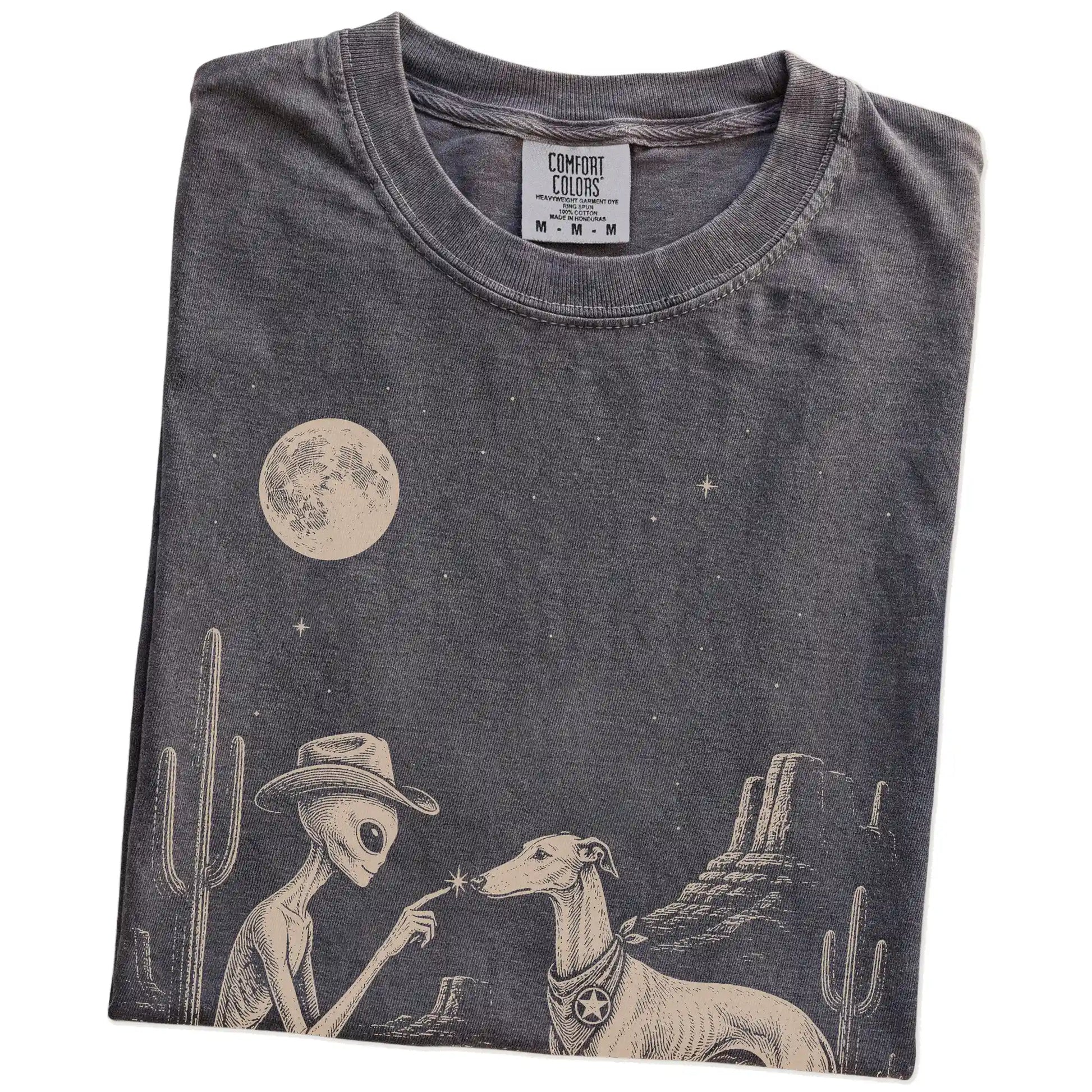 Folded Pepper Comfort Colors® T-shirt showing cowboy alien fist-bumping a greyhound beneath the moon