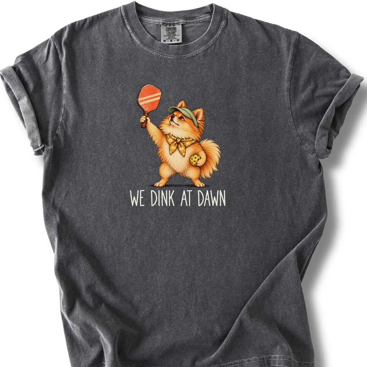 Pomeranian with a pickleball paddle and ball on a Pepper Comfort Colors® T-shirt with We Dink at Dawn text