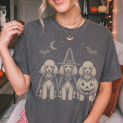 Model wearing a Pepper Comfort Colors® T-shirt showing three Poodle dogs dressed for Halloween beneath bats and a crescent mo