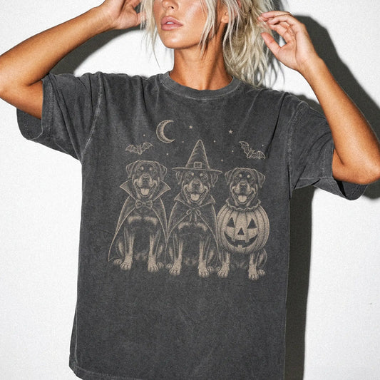 Model wearing a Pepper Comfort Colors® T-shirt showing three Rottweiler dogs dressed for Halloween beneath bats and a crescen