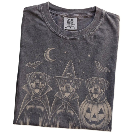 Folded Pepper Comfort Colors® T-shirt showing three Rottweiler dogs dressed for Halloween beneath bats and a crescent moon