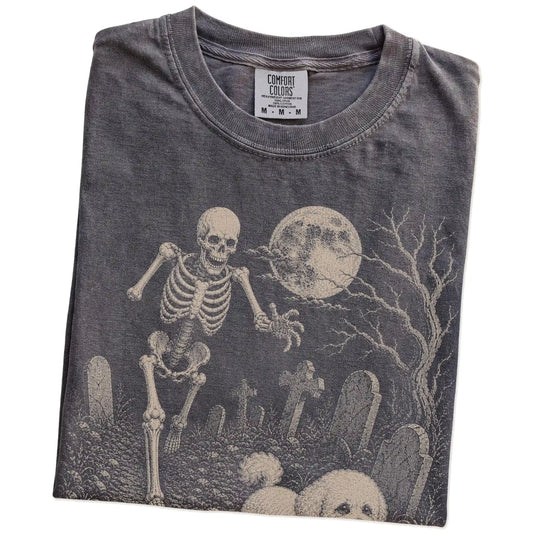 Folded Pepper Comfort Colors® T-shirt showing Bichon Frise running through a moonlit graveyard with a chasing skeleton