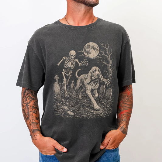 Model wearing a Pepper Comfort Colors® T-shirt showing Bloodhound running through a moonlit graveyard with a chasing skeleton