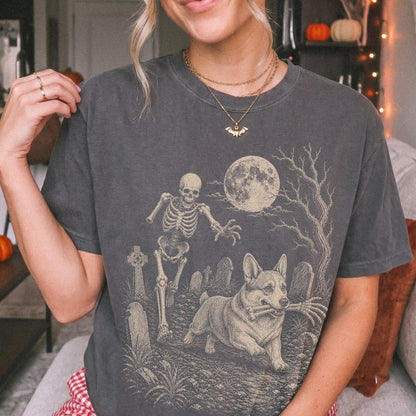 Model wearing a Pepper Comfort Colors® T-shirt showing Corgi running through a moonlit graveyard with a chasing skeleton