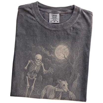 Folded Pepper Comfort Colors® T-shirt showing Greyhound running through a moonlit graveyard with a chasing skeleton