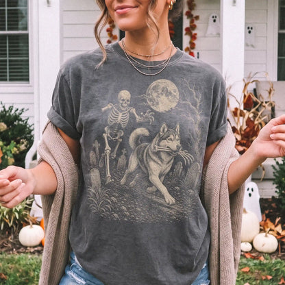 Model wearing a Pepper Comfort Colors® T-shirt showing Husky running through a moonlit graveyard with a chasing skeleton