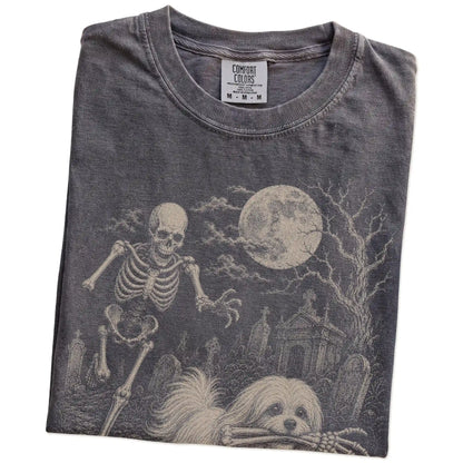 Folded Pepper Comfort Colors® T-shirt showing Maltese running through a moonlit graveyard with a chasing skeleton