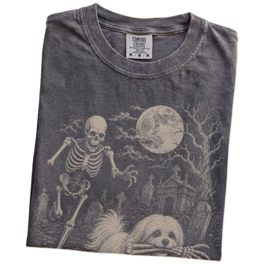 Folded Pepper Comfort Colors® T-shirt showing Maltese running through a moonlit graveyard with a chasing skeleton