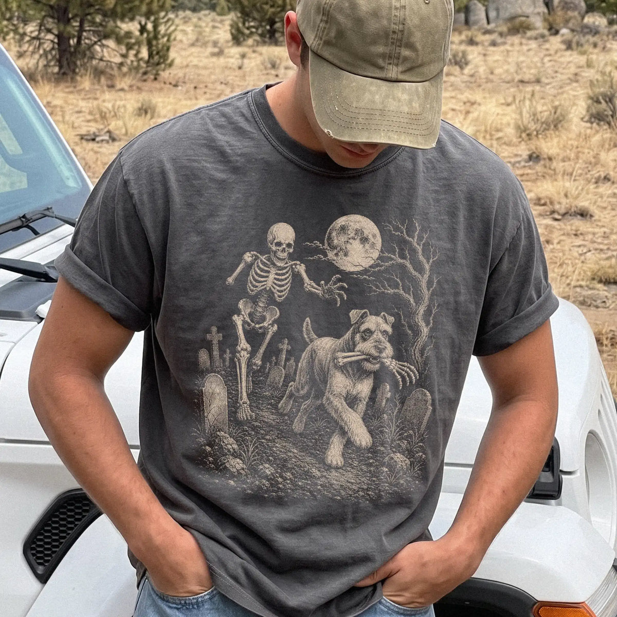 Model wearing a Pepper Comfort Colors® T-shirt showing Schnauzer running through a moonlit graveyard with a chasing skeleton
