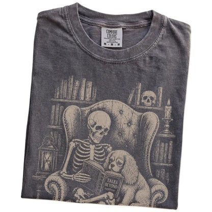 Folded Pepper Comfort Colors® T-shirt showing skeleton reading in a library beside a Cavalier King Charles Spaniel