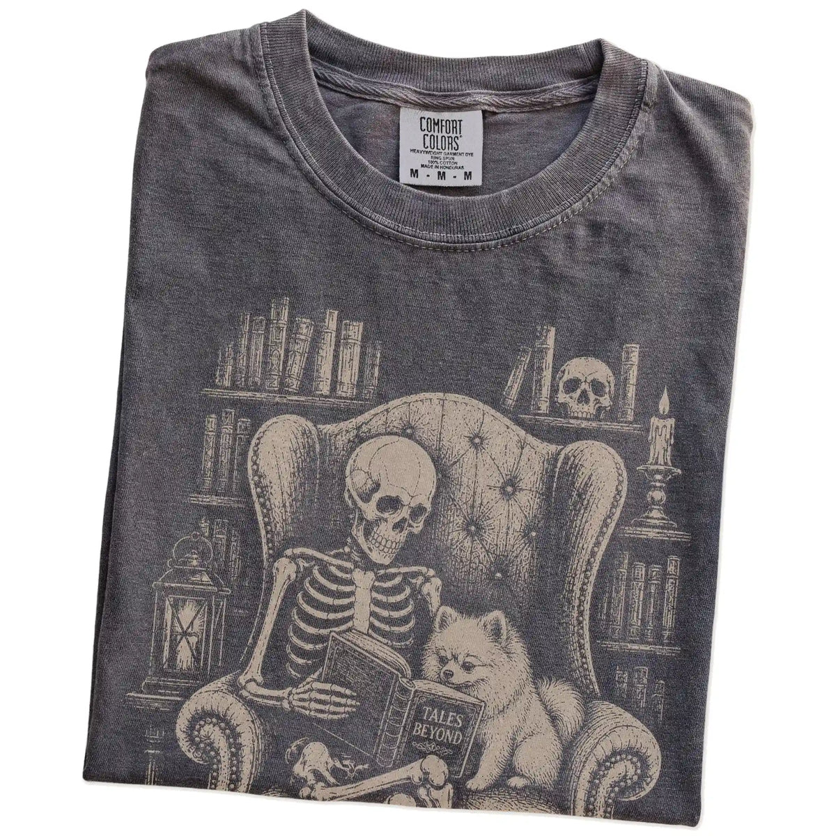 Folded Pepper Comfort Colors® T-shirt showing skeleton reading in a library beside a Pomeranian